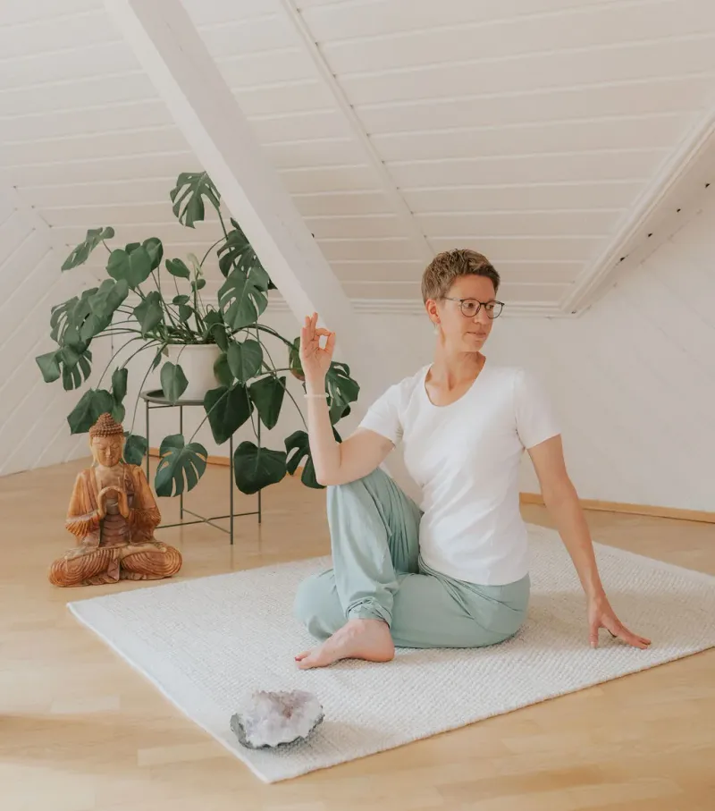 Yogini im Drehsitz auf einer Matte im Yogaraum unter dem Dach, daneben eine Monstera und eine Buddha-Figur