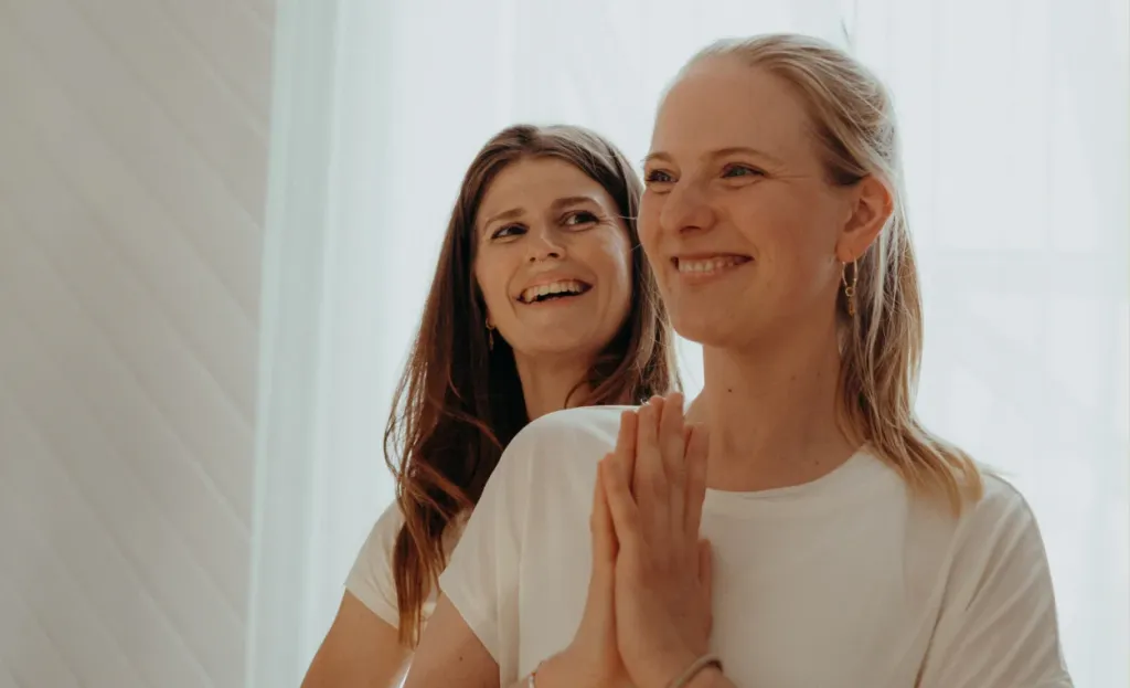 Zwei lachende Frauen mit Händen vor der Brust in einer Yogaklasse